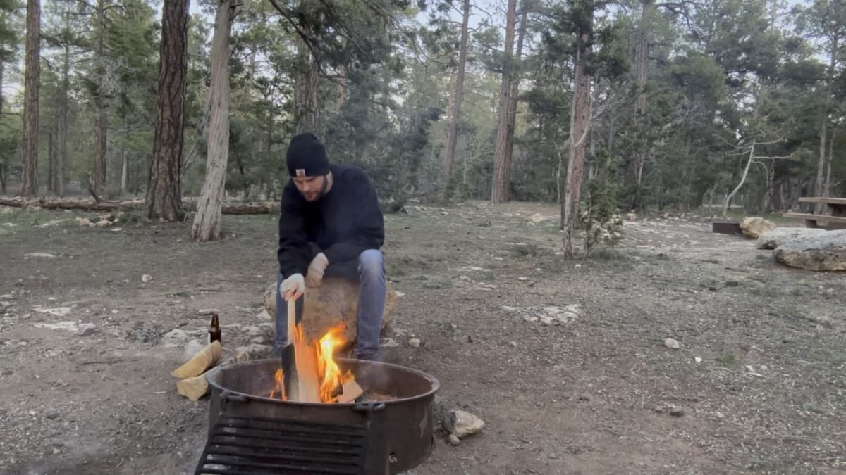 camper-tending-fire-mather-campground-grand-canyon A camper wearing a black beanie and jacket tending a campfire at Mather Campground in Grand Canyon National Park. Tall pine trees surround the campsite, and a picnic table is in the background.