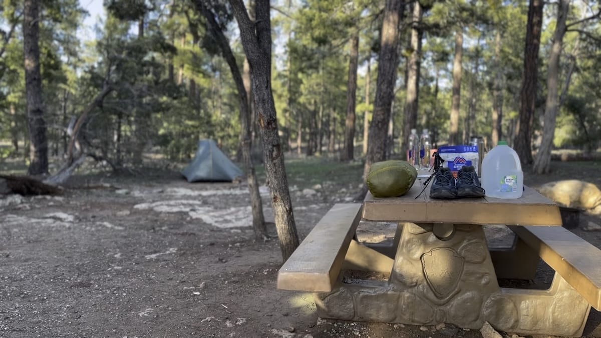 campsite-gear-picnic-table-mather-grand-canyon Camping gear, including hiking shoes, a stuff sack, and a water jug, on a picnic table at Mather Campground in Grand Canyon National Park. A tent is pitched in the background.
