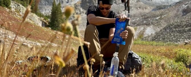 Hiker filtering water with a Sawyer Squeeze sitting on his backpack in a meadow.