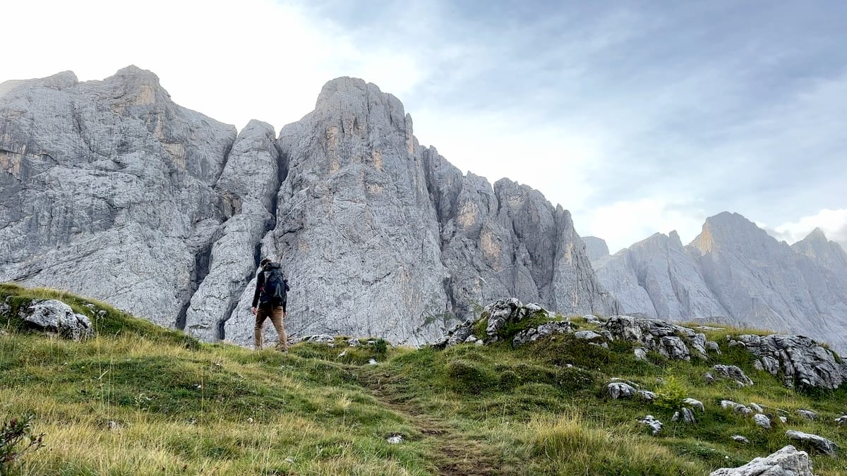 Alta Via 2 hiker over the ridge with massive peaks in the background and green grass in the foreground.