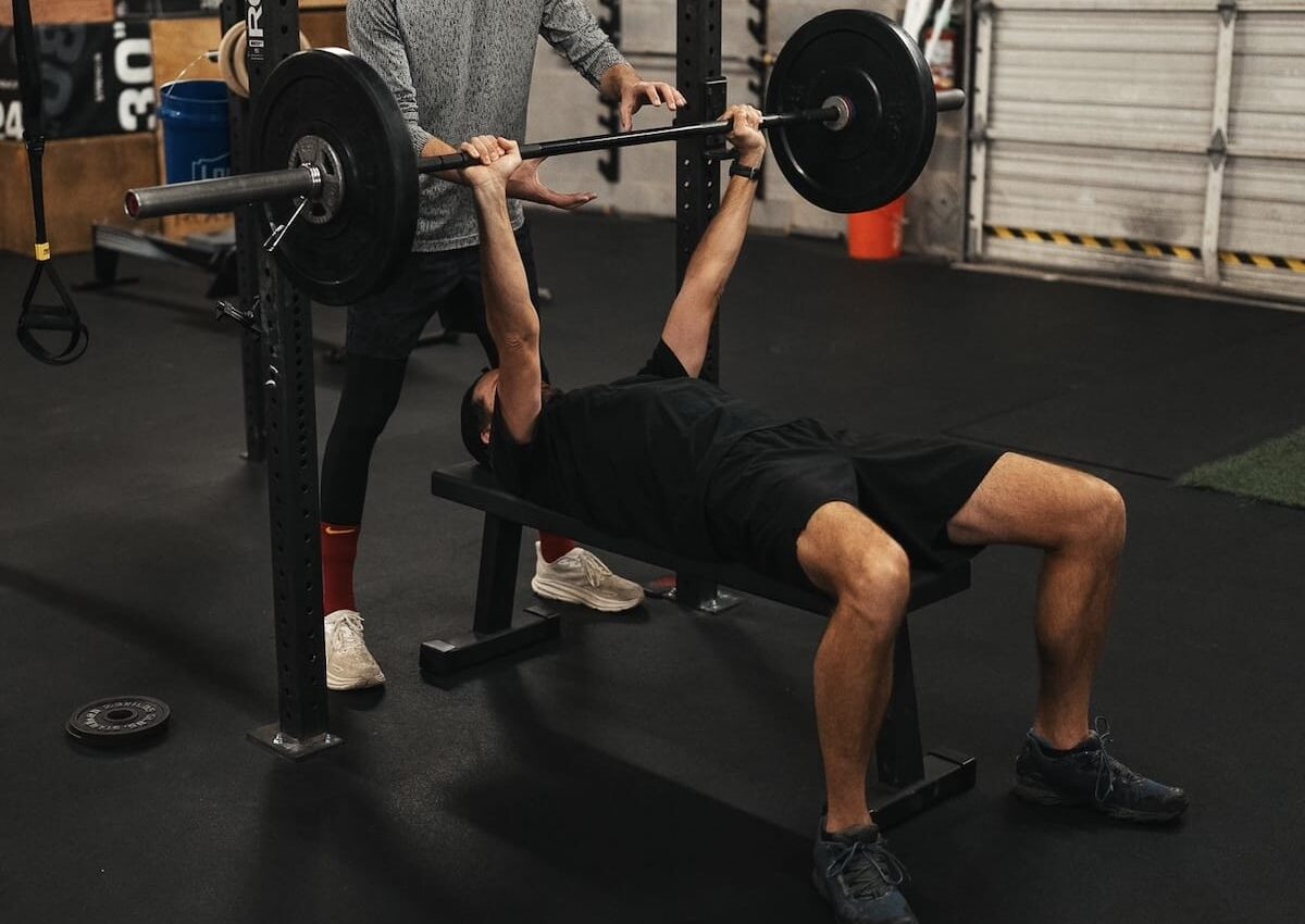 Man lifting barbell during a bench press exercise, with trainer spotting.
