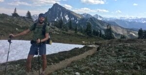 PCT thru hiker posing with trekking poles with large snow patched mountains in the background.