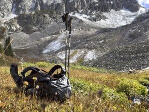 An Osprey Exos backpack laying in a grassy field with trekking poles standing up next to it and a mountain scene in the background.