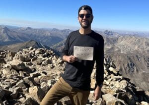 Hiker on Mt Elbert summit holding up elevation sign on a clear day.