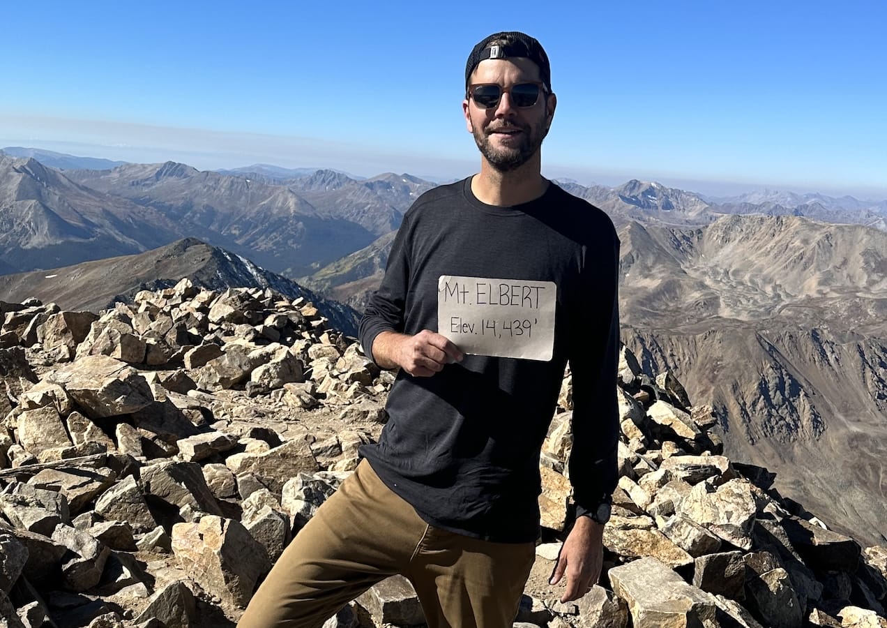 Hiker on Mt Elbert summit holding up elevation sign on a clear day.