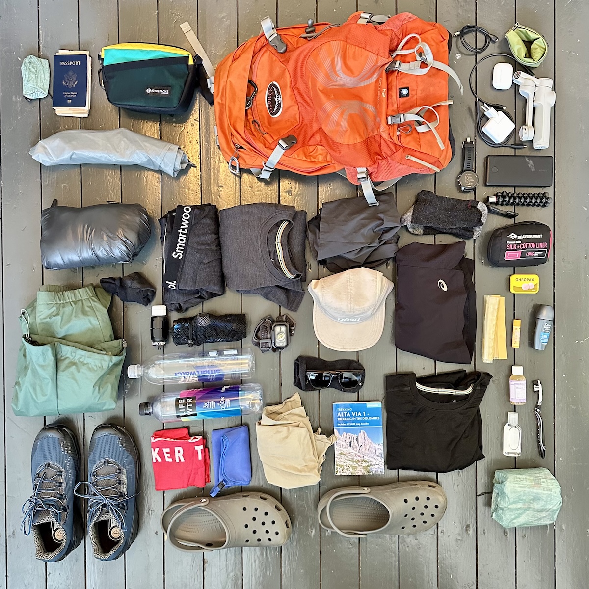 Complete Alta Via 1 gear list – backpack, hiking shoes, clothing layers, rain jacket, Crocs, trekking guidebook, passport, water bottles, and electronics laid out for Dolomites trek.