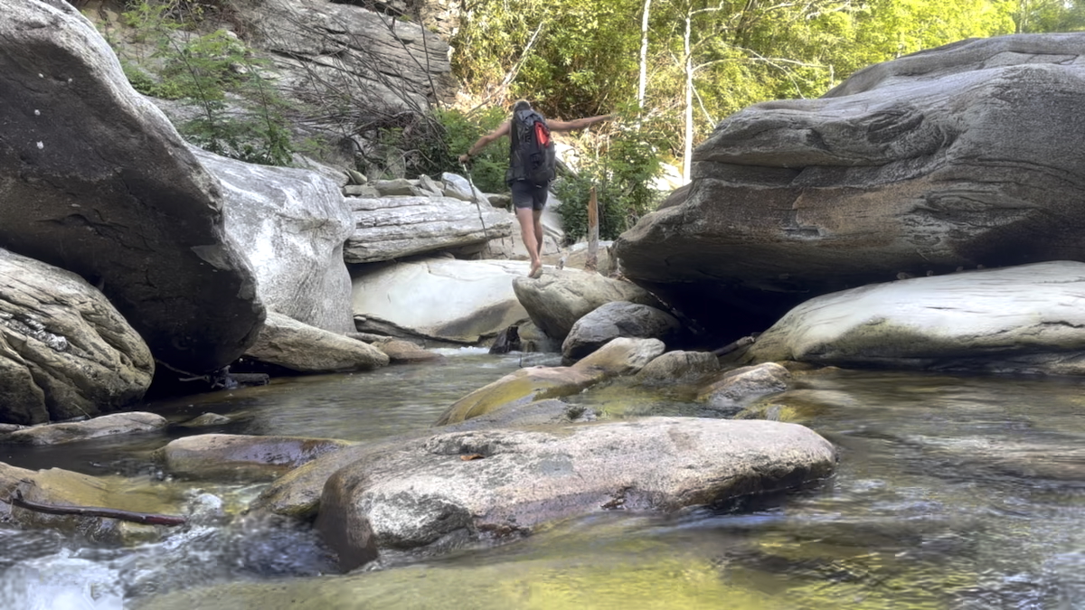 Hiker rock-hopping across the Linville River beneath forested gorge walls.