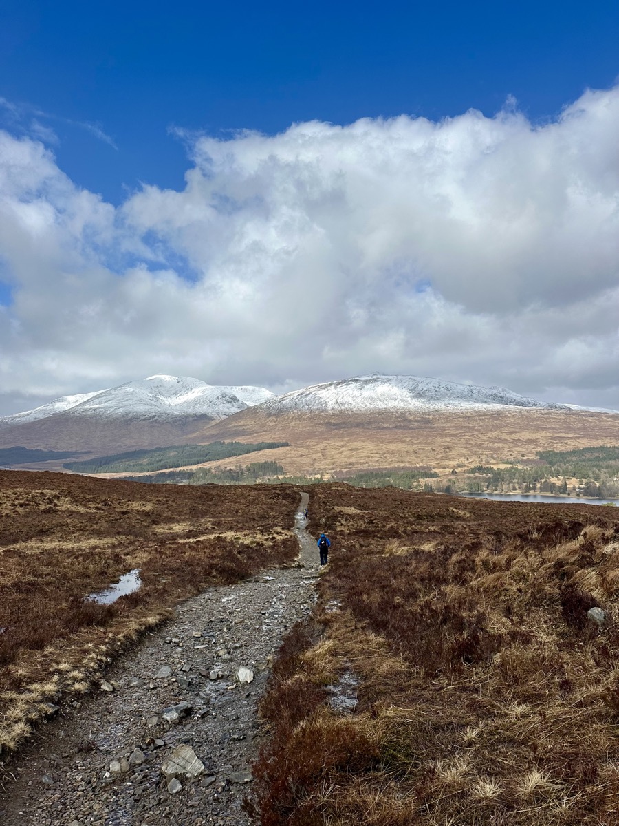 Hiker on the West Highland Way trail across Rannoch Moor with snow-capped mountains and Loch Tulla behind