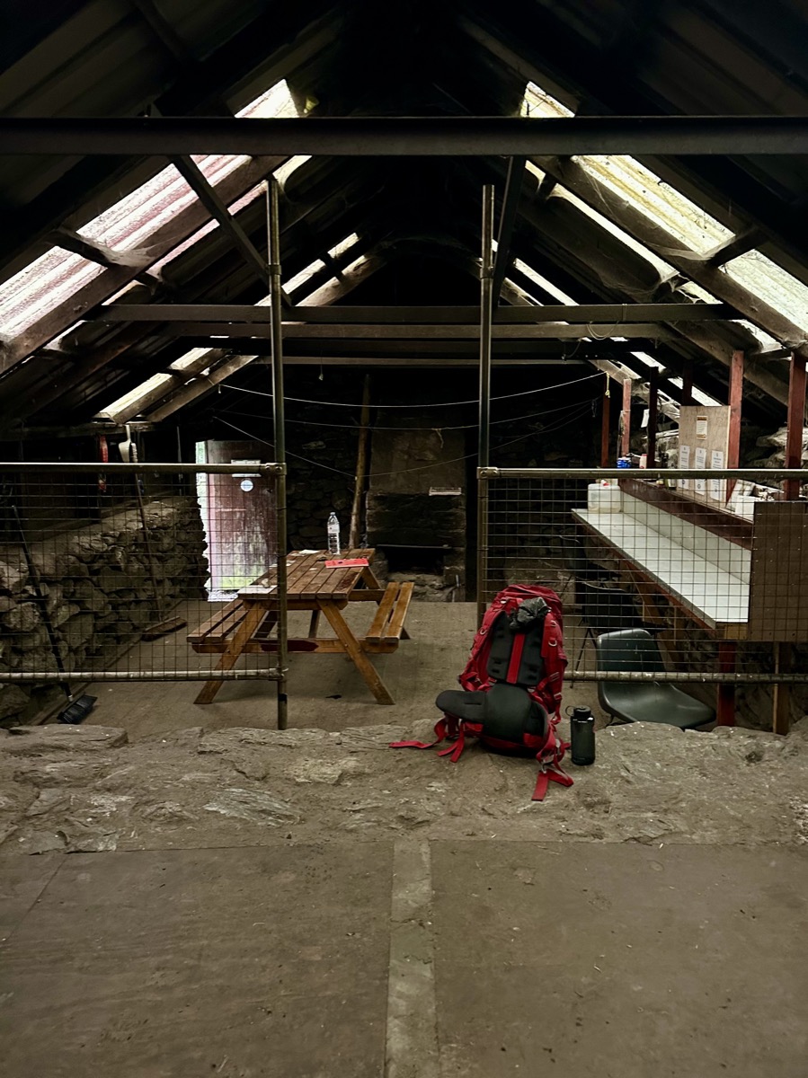 Wide interior view of Rowchoish Bothy on the West Highland Way showing sleeping platforms and hiker's pack