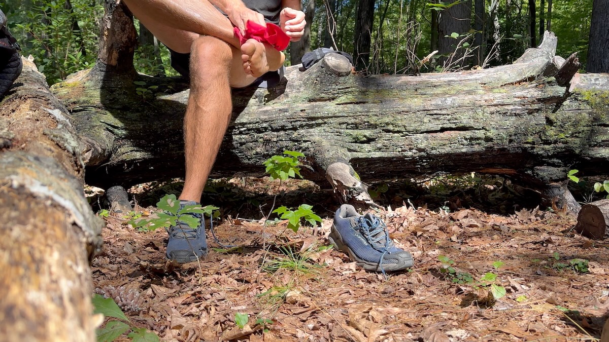 hiker-cleaning-foot-bandanna-trail-break Hiker wiping foot with a bandanna during a trail break seated on a log in the woods.