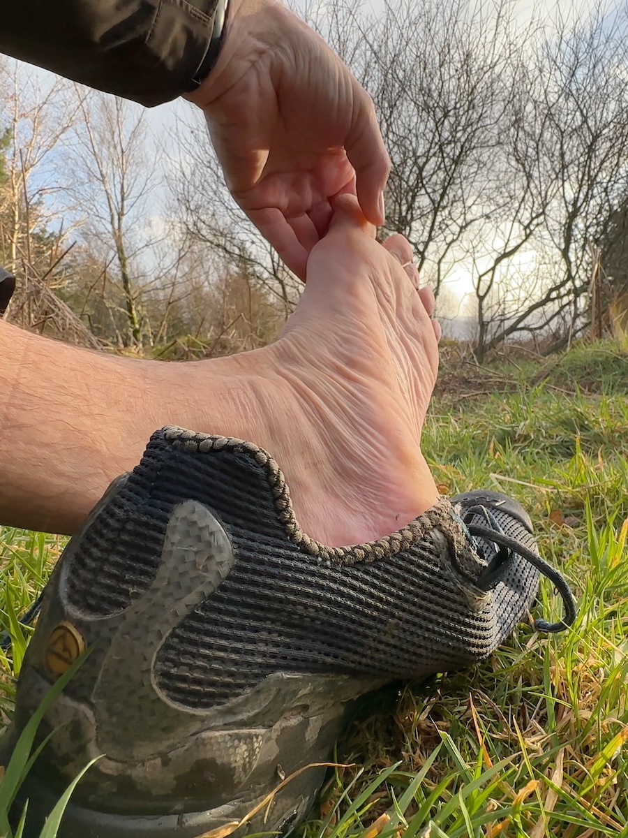 hiker-checking-bare-foot-rest-break-grass Hiker checking bare foot during a rest break on grass with trail runners nearby.